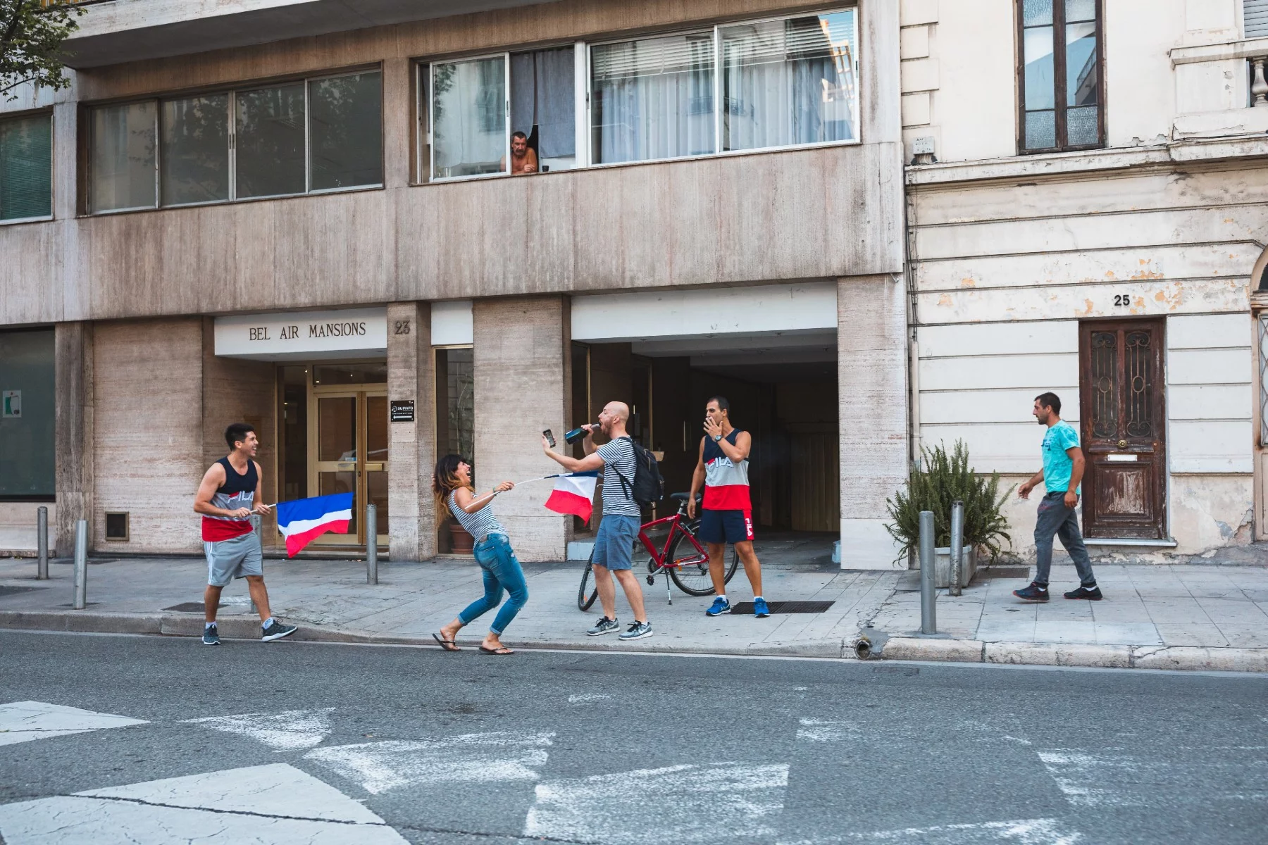 Bir apartmanın önündeki kaldırımda Fransız bayrakları sallayan ve kutlama yapan insanlar.