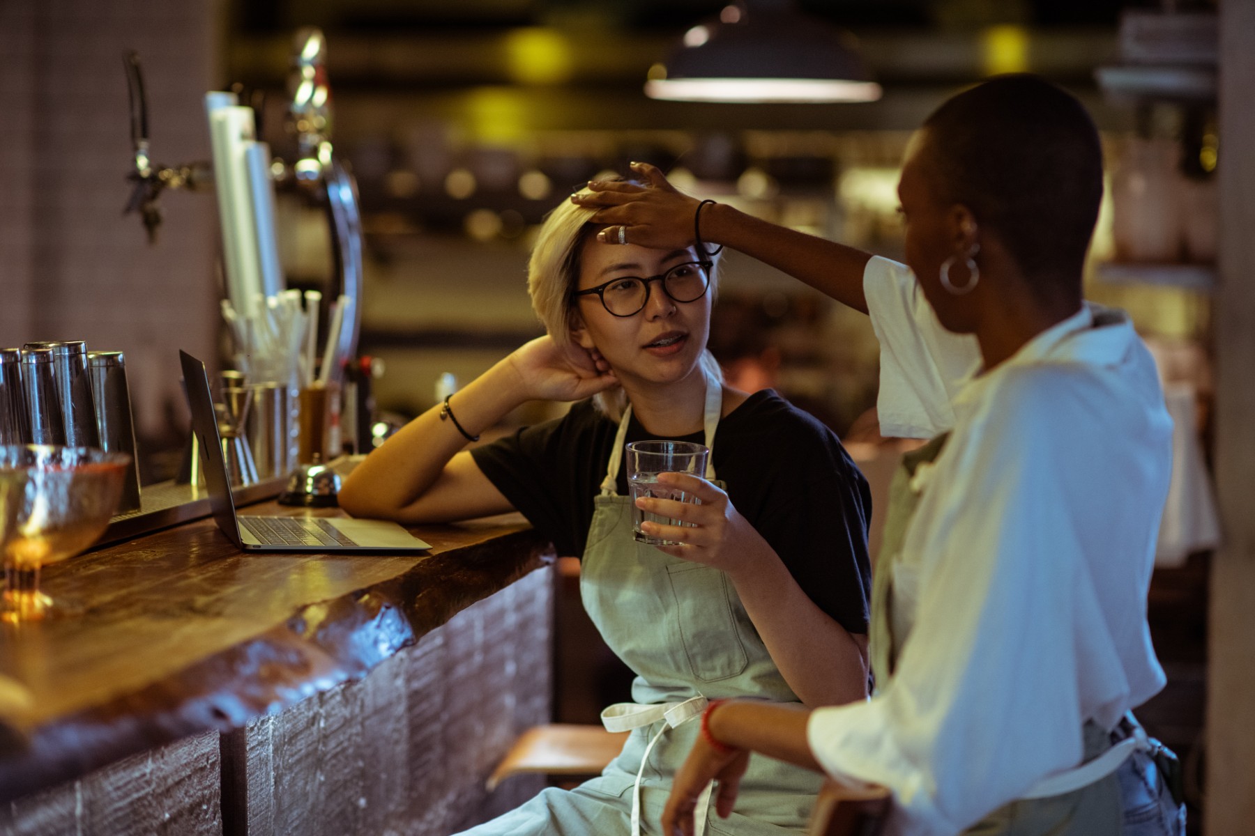 Bir restoranda barda oturan başka bir kadın işçinin alnını okşayan kadın işçi.