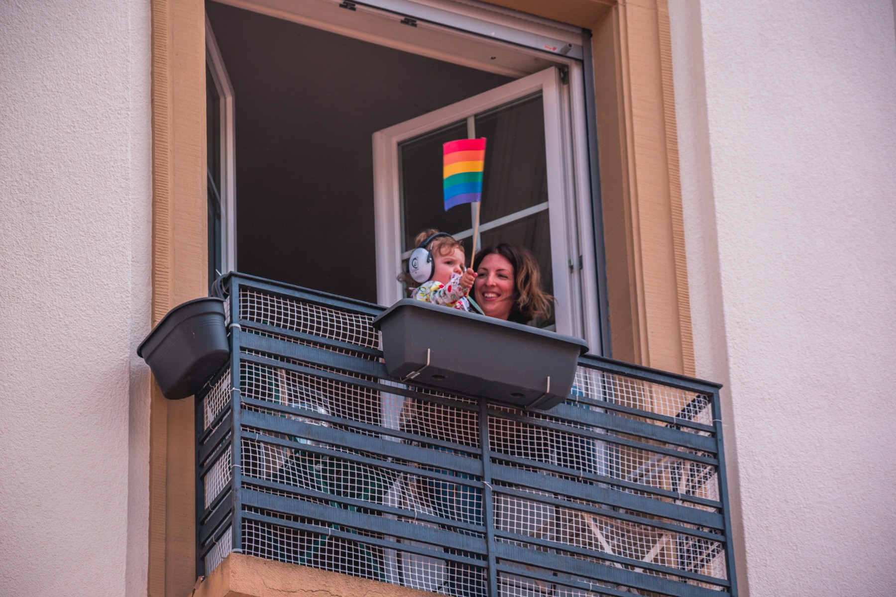 Anne ve bebek balkonda duruyor.  Anneleri onlara hayranlıkla bakarken bebek bir gökkuşağı bayrağı tutuyor.