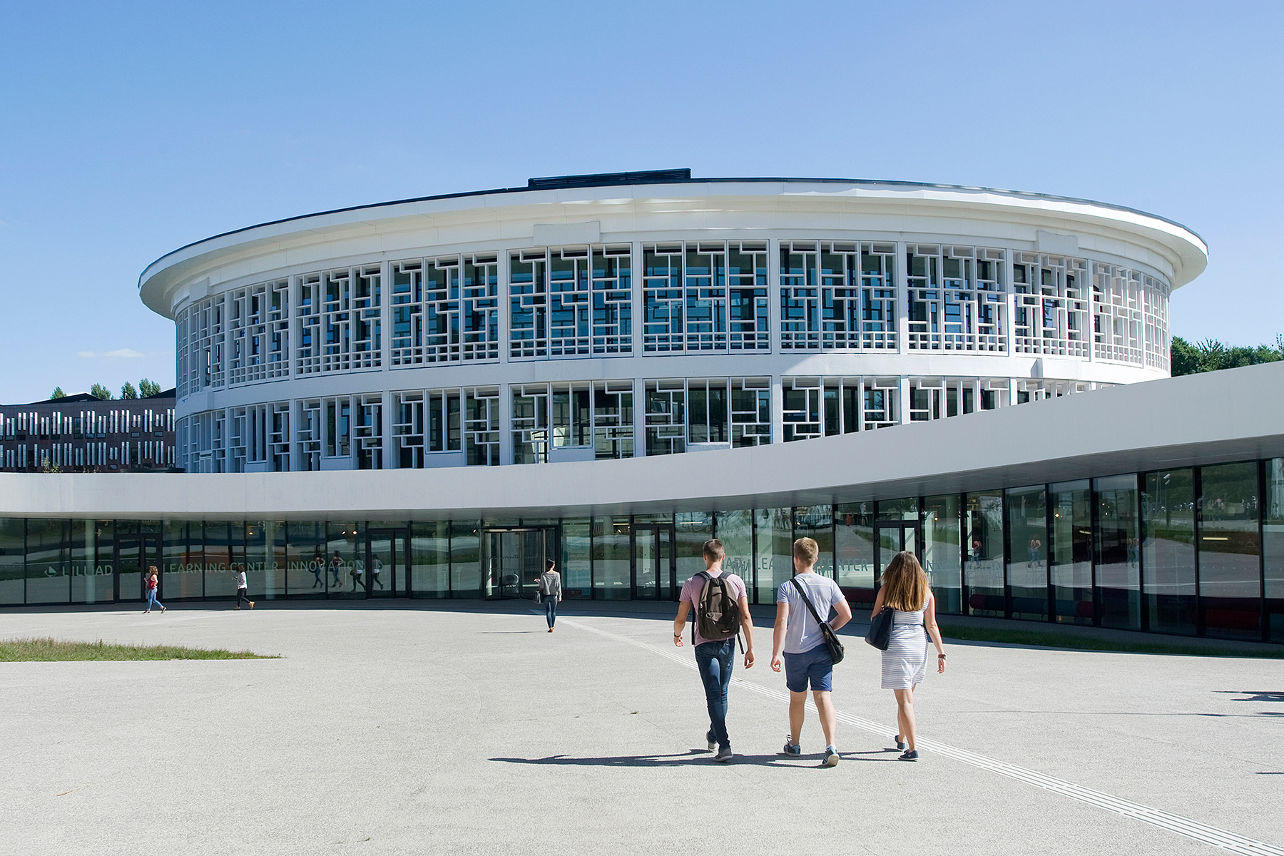 Güneşli bir günde büyük, beyaz, silindirik, modern bir üniversite binasının önünde yürüyen öğrenciler.